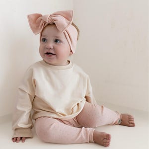 May include: A baby wearing a light pink headband with a large bow, a cream-colored long-sleeved shirt, and pink leggings with a small dot pattern. The baby is sitting and looking to the side.