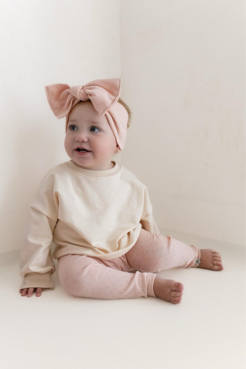 May include: A baby wearing a light pink headband with a large bow, a cream-colored long-sleeved shirt, and pink leggings with a small dot pattern. The baby is sitting and looking to the side.