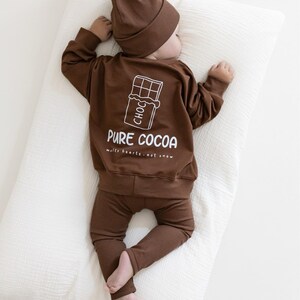 Puede incluir: Un conjunto de ropa de beb&eacute; marr&oacute;n, que incluye una camiseta de manga larga, pantalones y un gorro. La camiseta presenta un gr&aacute;fico blanco de una barra de chocolate y las palabras "PURE COCOA" con la frase "melts hearts - not snow". El beb&eacute; est&aacute; acostado sobre una almohada blanca.