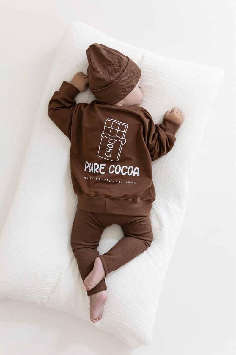 Puede incluir: Un conjunto de ropa de beb&eacute; marr&oacute;n, que incluye una camiseta de manga larga, pantalones y un gorro. La camiseta presenta un gr&aacute;fico blanco de una barra de chocolate y las palabras "PURE COCOA" con la frase "melts hearts - not snow". El beb&eacute; est&aacute; acostado sobre una almohada blanca.