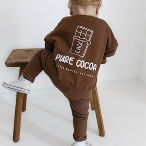 Puede incluir: Conjunto marr&oacute;n de sudadera y pantal&oacute;n para ni&ntilde;o. La sudadera presenta un gr&aacute;fico blanco de una tableta de chocolate y las palabras "PURE COCOA" con la frase "melts hearts - not snow". El ni&ntilde;o lleva zapatillas blancas.
