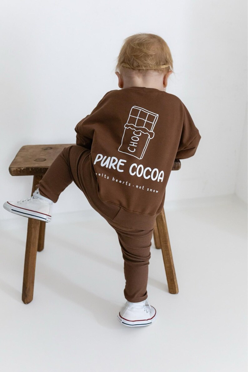 Puede incluir: Conjunto marr&oacute;n de sudadera y pantal&oacute;n para ni&ntilde;o. La sudadera presenta un gr&aacute;fico blanco de una tableta de chocolate y las palabras "PURE COCOA" con la frase "melts hearts - not snow". El ni&ntilde;o lleva zapatillas blancas.