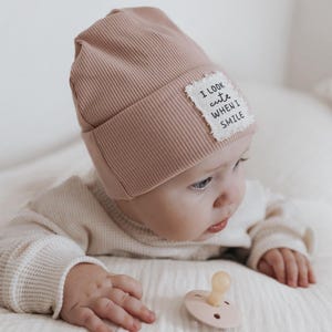 Könnte beinhalten: Ein Baby trägt eine rosa gerippte Mütze mit einem weißen Patch, auf dem "Ich sehe süß aus, wenn ich lächle" steht. Das Baby liegt auf dem Bauch und hat einen Schnuller im Mund.