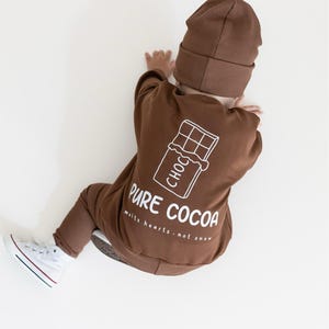 Puede incluir: Conjunto de beb&eacute; marr&oacute;n con gorro a juego. El conjunto presenta un gr&aacute;fico blanco de una tableta de chocolate con las palabras "PURE COCOA" y "melts hearts - not snow". El beb&eacute; lleva zapatillas blancas con rayas rojas y azules.