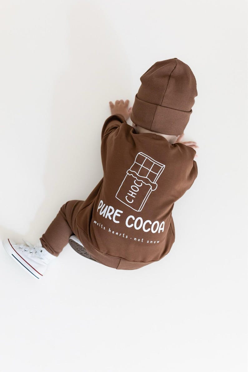Puede incluir: Conjunto de beb&eacute; marr&oacute;n con gorro a juego. El conjunto presenta un gr&aacute;fico blanco de una tableta de chocolate con las palabras "PURE COCOA" y "melts hearts - not snow". El beb&eacute; lleva zapatillas blancas con rayas rojas y azules.