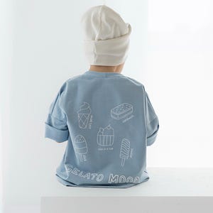 Op de afbeelding: Een kind draagt een lichtblauw T-shirt en een witte muts. De achterkant van het shirt heeft witte lijntekeningen van ijsjes en de woorden "GELATO MOOD".