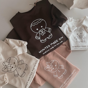 Könnte beinhalten: Verschiedene Sweatshirts in Braun-, Creme- und Rosatönen, jeweils mit einem weißen Lebkuchenmann-Motiv. Die Textaufdrucke lauten entweder "WINTER MODE ON" oder "SWEET WINTER MOMENTS".