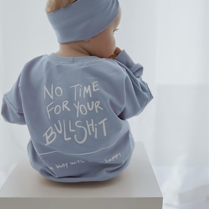Könnte beinhalten: Ein Baby trägt einen hellblauen Sweatshirt und ein passendes Stirnband mit Schleife. Auf der Rückseite des Sweatshirts steht in Weiß der Text "NO TIME FOR YOUR BULLSHIT". Der Text "busy with happy" ist ebenfalls sichtbar.