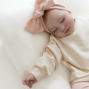 Puede incluir: Un beb&eacute; dormido con una diadema de color melocot&oacute;n claro con un lazo grande. El beb&eacute; viste una camisa de manga larga color crema y pantalones rosas claros estampados, descansando sobre una almohada color crema.