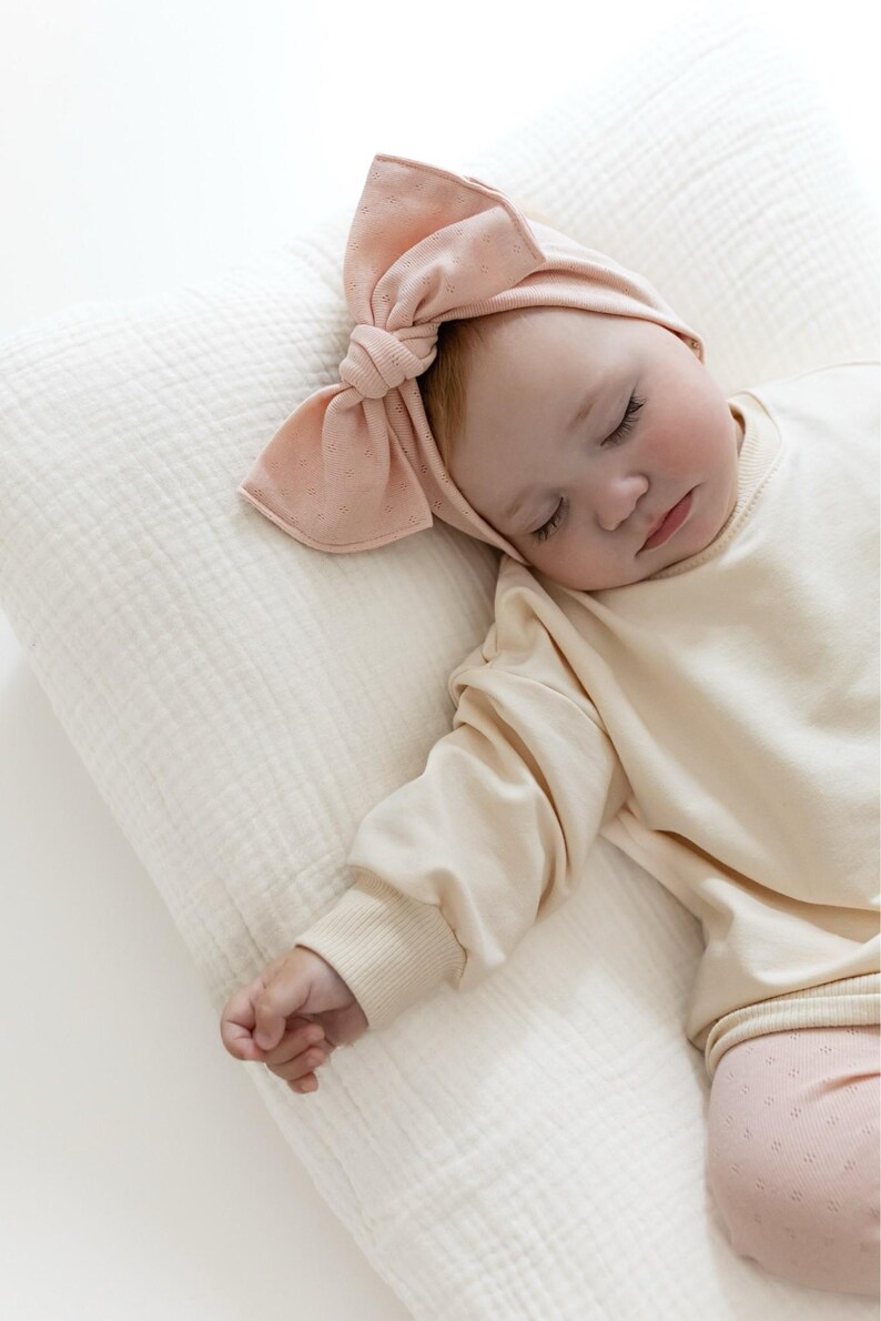 Puede incluir: Un beb&eacute; dormido con una diadema de color melocot&oacute;n claro con un lazo grande. El beb&eacute; viste una camisa de manga larga color crema y pantalones rosas claros estampados, descansando sobre una almohada color crema.