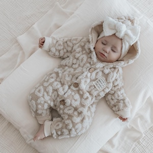 Könnte beinhalten: Ein Baby trägt einen cremefarbenen Kapuzenoverall mit Leopardenmuster. Der Overall hat einen Knopfverschluss und einen Aufnäher mit dem Text "Dream Big Little One". Das Baby trägt ein weißes Stirnband mit einer großen Schleife.