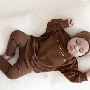 Puede incluir: Un beb&eacute; dormido con un conjunto de punto acanalado marr&oacute;n, que incluye una parte superior de manga larga, pantalones y un gorro. La parte superior tiene el texto "choco state of mind". El beb&eacute; est&aacute; acostado sobre una almohada blanca.