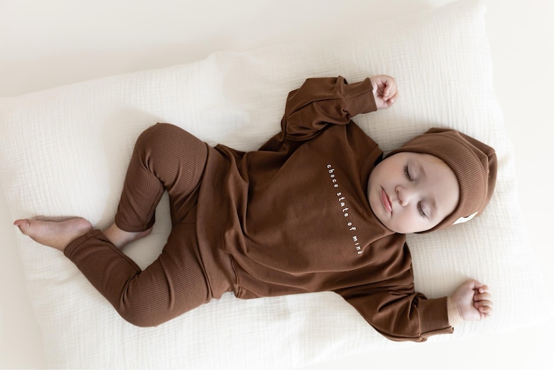 Puede incluir: Un beb&eacute; dormido con un conjunto de punto acanalado marr&oacute;n, que incluye una parte superior de manga larga, pantalones y un gorro. La parte superior tiene el texto "choco state of mind". El beb&eacute; est&aacute; acostado sobre una almohada blanca.