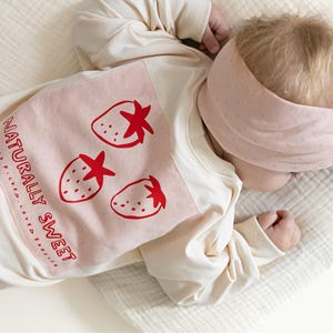 May include: A baby wearing a cream-colored sweatshirt with a pink strawberry design and the words "NATURALLY SWEET." The baby also wears a matching pink headband. The outfit is on a white surface.