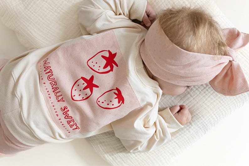 May include: A baby wearing a cream-colored sweatshirt with a pink strawberry design and the words "NATURALLY SWEET." The baby also wears a matching pink headband. The outfit is on a white surface.