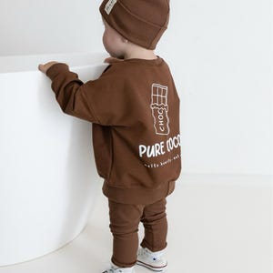 Puede incluir: Un conjunto de beb&eacute; marr&oacute;n, que incluye gorro, sudadera y pantal&oacute;n. La sudadera presenta un gr&aacute;fico blanco de una tableta de chocolate con las palabras "PURE COCO" impresas debajo. El conjunto se combina con zapatillas blancas.
