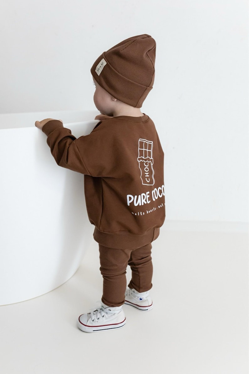 Puede incluir: Un conjunto de beb&eacute; marr&oacute;n, que incluye gorro, sudadera y pantal&oacute;n. La sudadera presenta un gr&aacute;fico blanco de una tableta de chocolate con las palabras "PURE COCO" impresas debajo. El conjunto se combina con zapatillas blancas.