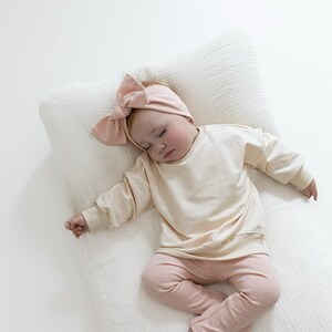 May include: A sleeping baby wearing a light beige sweatshirt, pink leggings, and a matching bow headband. The baby is lying on a white, textured pillow. The outfit is soft and comfortable, perfect for a newborn.