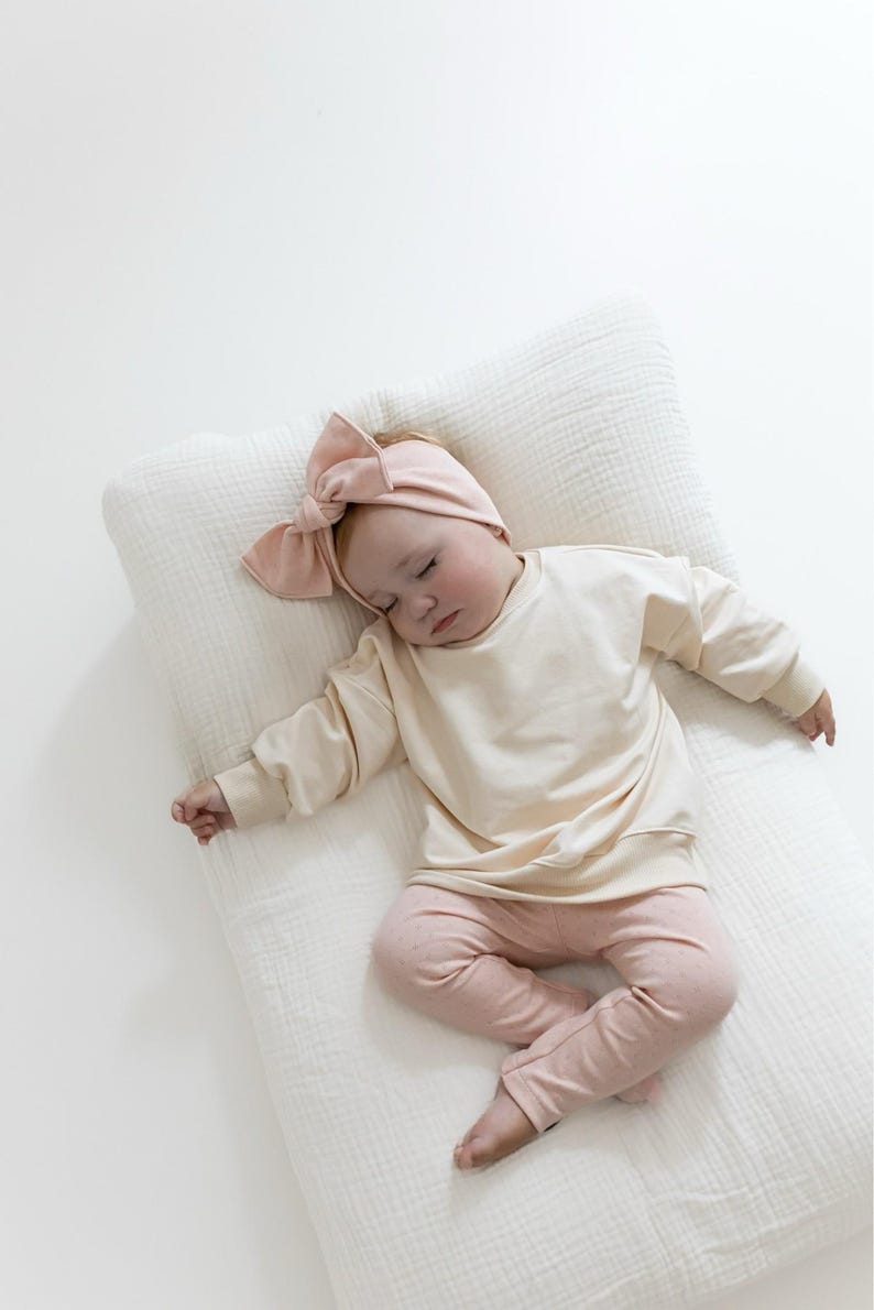 May include: A sleeping baby wearing a light beige sweatshirt, pink leggings, and a matching bow headband. The baby is lying on a white, textured pillow. The outfit is soft and comfortable, perfect for a newborn.