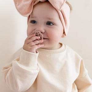 May include: A baby wearing a light pink bow headband and a matching pacifier. The baby is also wearing a cream-colored sweatshirt and pink patterned pants. The baby is smiling and looking to the side.