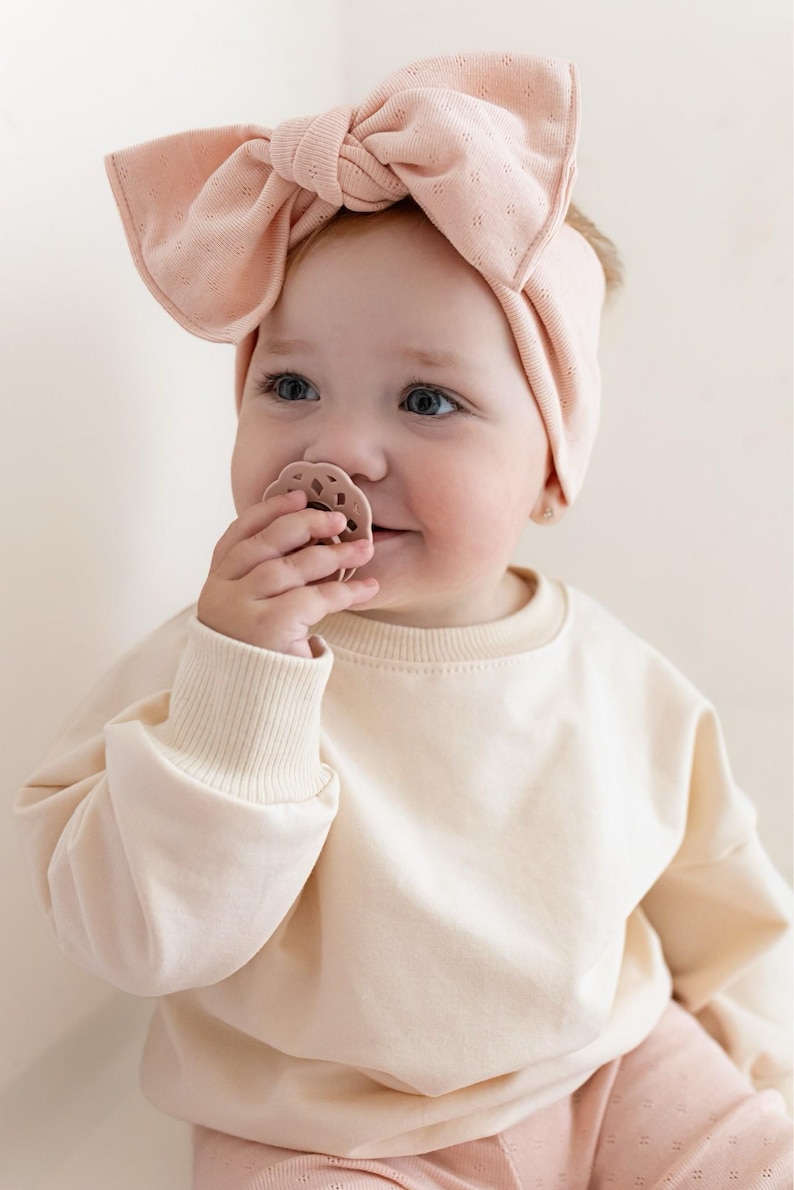 May include: A baby wearing a light pink bow headband and a matching pacifier. The baby is also wearing a cream-colored sweatshirt and pink patterned pants. The baby is smiling and looking to the side.