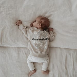 Könnte beinhalten: Ein Baby in einem beigefarbenen Sweatshirt und einer weißen Hose liegt auf einem cremefarbenen Bett. Auf dem Sweatshirt stehen die Worte "LOVE, I love my daddy, LOVE, LOVE, LOVE" in Weiß. Das Baby liegt auf dem Bauch.