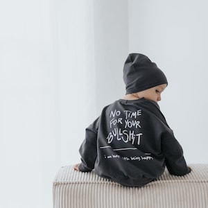 Könnte beinhalten: Ein Kind trägt einen dunkelgrauen Pullover und eine Mütze. Der Pullover hat den weißen Text "NO TIME FOR YOUR BULLSHIT". Das Kind sitzt auf einer hellen, strukturierten Bank.