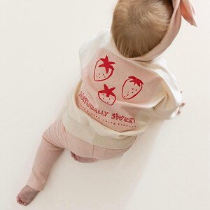 May include: A baby wearing a light pink headband, sweatshirt, and pants. The white sweatshirt has a pink panel with three red strawberries and the text "NATURALLY SWEET". The baby is sitting on a white surface.