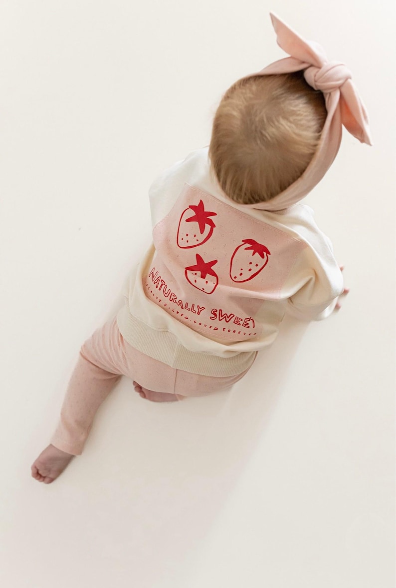 May include: A baby wearing a light pink headband, sweatshirt, and pants. The white sweatshirt has a pink panel with three red strawberries and the text "NATURALLY SWEET". The baby is sitting on a white surface.