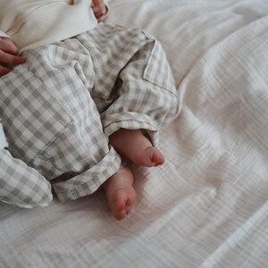 Könnte beinhalten: Ein Baby in einem cremefarbenen Pullover und einer grau-weiß karierten Hose. Die Füße des Babys sind sichtbar und liegen auf einer weißen, strukturierten Decke. Ein Stofftier ist links teilweise zu sehen.