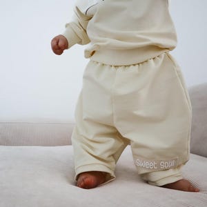 Könnte beinhalten: Ein cremefarbenes zweiteiliges Set bestehend aus einem langärmeligen Sweatshirt und einer locker sitzenden Hose. Die Hose hat einen rechteckigen Aufnäher am unteren linken Bein mit dem Text "Sweet soul", der in einer hellen Schreibschrift gestickt ist.