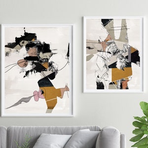May include: Two abstract art prints in white frames, featuring black, white, gray, and gold geometric shapes and lines. The prints are hanging on a white wall above a gray sofa with two pillows.