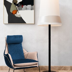 May include: A blue velvet armchair with a beige cushion sits in front of a white wall with a framed abstract painting and a tall black floor lamp with a white lampshade.