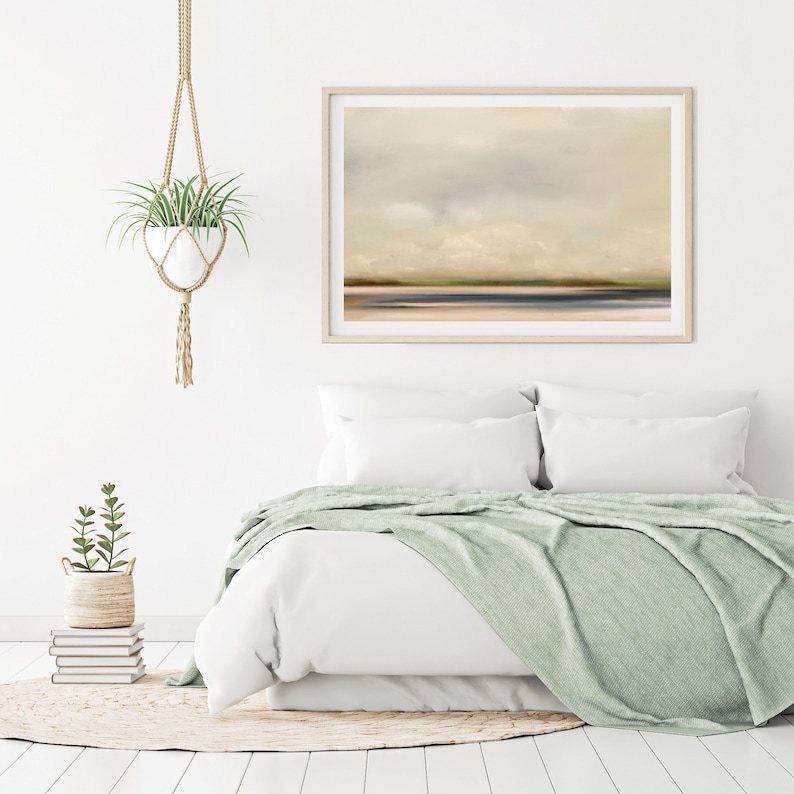 May include: A white bed with a light green throw blanket and two white pillows. A framed abstract painting of a landscape with a cloudy sky hangs above the bed. A potted plant hangs from the ceiling in the upper left corner of the image.