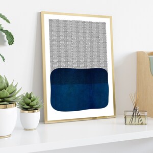 May include: Abstract art print in a gold frame. The artwork features a textured black and white pattern at the top and a dark blue, rectangular shape at the bottom. The print is displayed on a white shelf with decorative plants.
