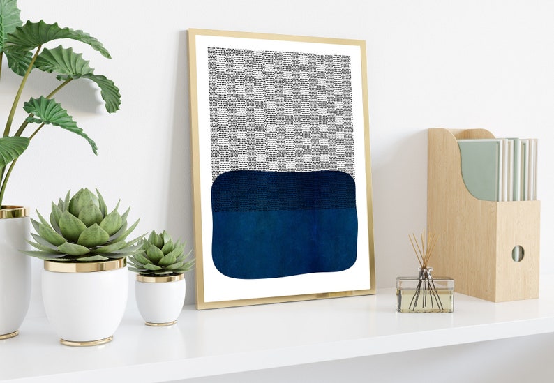 May include: Abstract art print in a gold frame. The artwork features a textured black and white pattern at the top and a dark blue, rectangular shape at the bottom. The print is displayed on a white shelf with decorative plants.