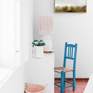 Op de afbeelding: Een witte kamer met een blauwe stoel, een witte tafel met een plant en een lamp, en een ingelijst schilderij aan de muur. De vloer is betegeld met rood.