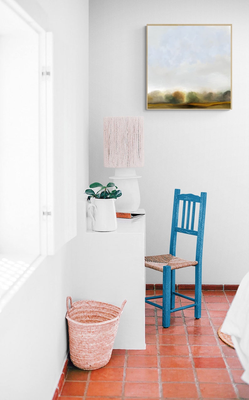 Op de afbeelding: Een witte kamer met een blauwe stoel, een witte tafel met een plant en een lamp, en een ingelijst schilderij aan de muur. De vloer is betegeld met rood.
