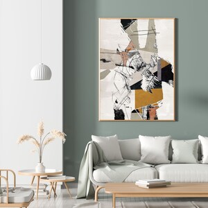 May include: Abstract art print in black, white, gray, and gold on a white background with a wood frame. The print is hanging on a sage green wall above a white sofa with a throw blanket.