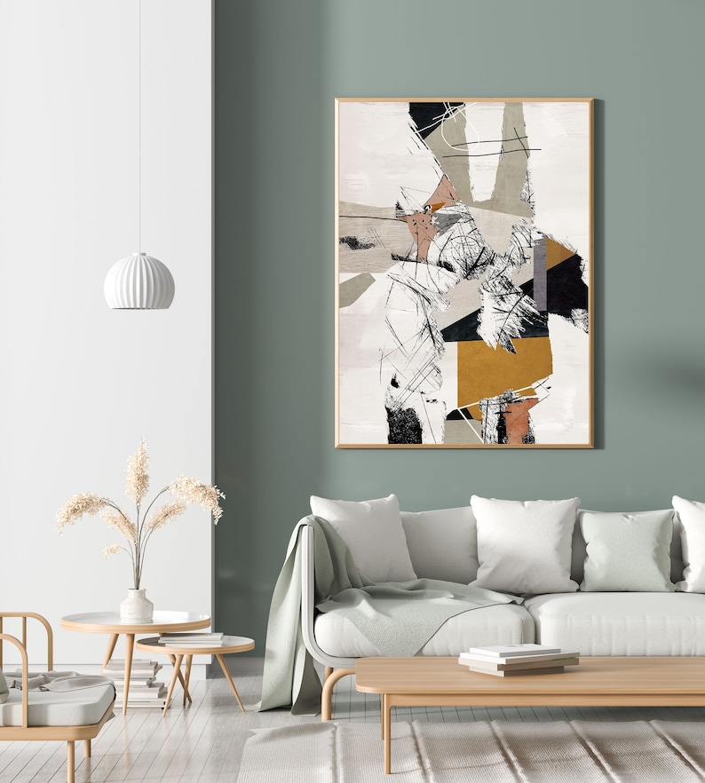 May include: Abstract art print in black, white, gray, and gold on a white background with a wood frame. The print is hanging on a sage green wall above a white sofa with a throw blanket.