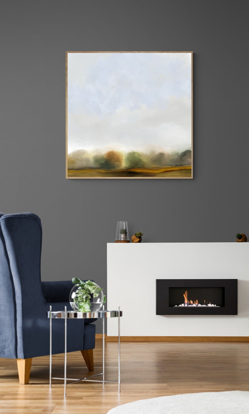 Op de afbeelding: Een ingelijst abstract landschapschilderij met een gedempte kleurenpalet van blauw, groen en bruin. Het schilderij toont een mistig veld met bomen in de verte. Het schilderij hangt aan een grijze muur boven een witte open haard met een zwarte vuurhaard.