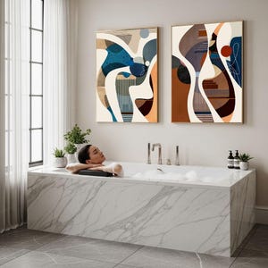 May include: A modern bathroom scene featuring a marble bathtub with a person relaxing in it. Two abstract art pieces hang on the wall. The artwork features a mix of blue, brown, and orange hues. Small potted plants and toiletries are visible.