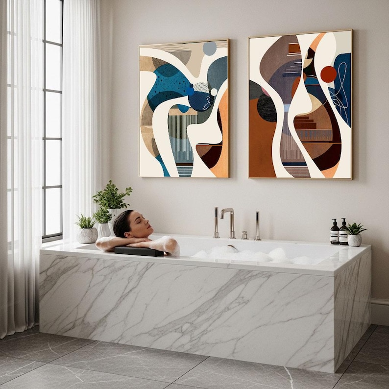 May include: A modern bathroom scene featuring a marble bathtub with a person relaxing in it. Two abstract art pieces hang on the wall. The artwork features a mix of blue, brown, and orange hues. Small potted plants and toiletries are visible.