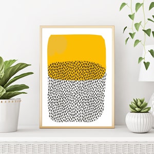 May include: Abstract art print in a gold frame. The artwork features a yellow top section, a middle section with black dots, and a bottom section with black dots. The print is displayed on a white shelf with plants and a lamp.