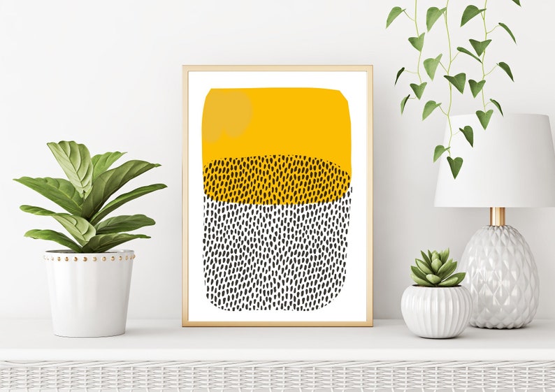May include: Abstract art print in a gold frame. The artwork features a yellow top section, a middle section with black dots, and a bottom section with black dots. The print is displayed on a white shelf with plants and a lamp.