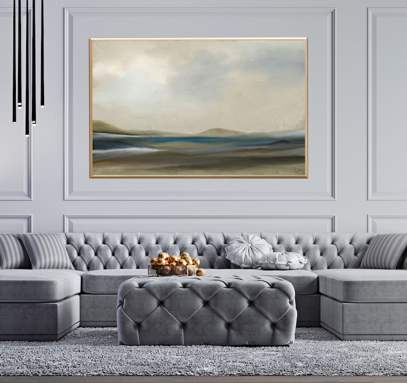 K&ouml;nnte beinhalten: Ein graues Ecksofa mit gesteppter Polsterung und ein grauer, gesteppter Ottoman vor einem gro&szlig;en abstrakten Landschaftsbild mit goldenem Rahmen. Das Bild zeigt einen blauen See mit braunen H&uuml;geln im Hintergrund.