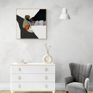 May include: A white dresser with three drawers and gold handles. The dresser has a white and gray abstract painting above it. A gray armchair is to the right of the dresser. A white lamp hangs from the ceiling above the dresser.