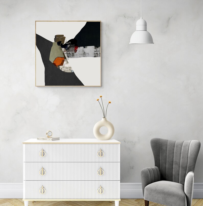 May include: A white dresser with three drawers and gold handles. The dresser has a white and gray abstract painting above it. A gray armchair is to the right of the dresser. A white lamp hangs from the ceiling above the dresser.