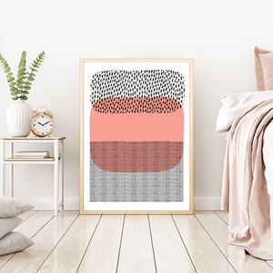 May include: Abstract art print in a gold frame. The artwork features a design with black, coral, and gray geometric shapes. The print is displayed in a bedroom setting, with a white bed and side table.