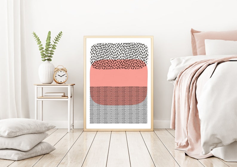May include: Abstract art print in a gold frame. The artwork features a design with black, coral, and gray geometric shapes. The print is displayed in a bedroom setting, with a white bed and side table.